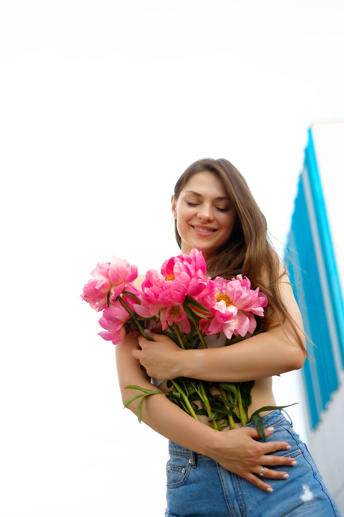 Eine junge kaukasische Frau mit einem Strauß leuchtender Farben, ein Mädchen in Jeanshosen im Sommer mit Blumenstrauß, eine Dame in Sommerkleidung gegen den Himmel - Foto, Bild