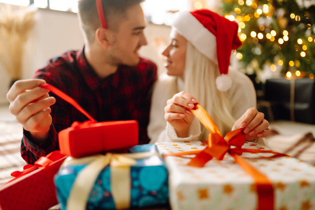 Fröhliche junge Männer und Frauen tauschen Weihnachtsgeschenke aus. Junges Paar öffnet Geschenkbox neben Weihnachtsbaum. Romantischer Tag. Winterurlaub. - Foto, Bild