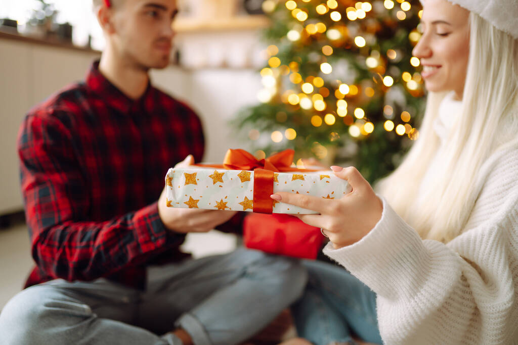 Веселый молодой мужчина и женщина Обмен рождественскими подарками. Молодая пара открывает подарочную коробку возле елки. Романтический день. Зимние каникулы. - Фото, изображение