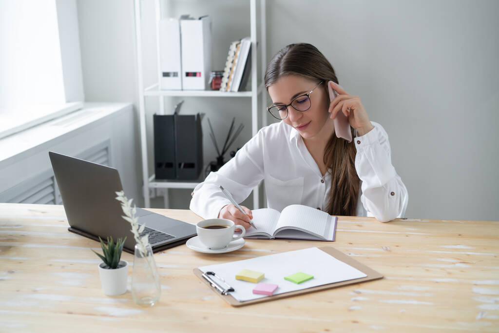 Junge Geschäftsfrau telefoniert, während sie zu Hause am Tisch sitzt, vor dem Laptop. Schreibt Informationen in Arbeitsnotizbuch. - Foto, Bild