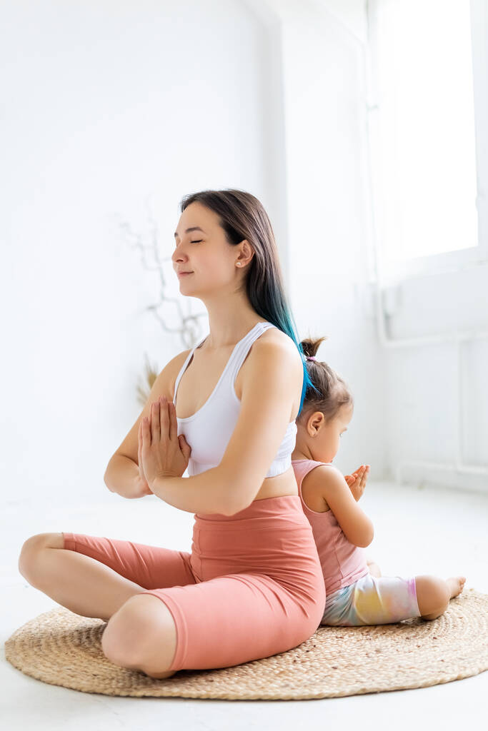 Young woman and little girl sit in lotus position back to back. Morning yoga practice. - Photo, Image