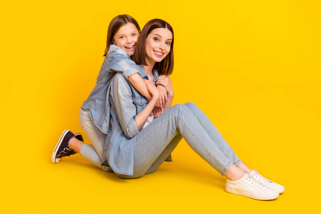 Fotografie rozkošné krásné sourozence oblečené džínové košile objímání sedací podlahy s úsměvem izolované žluté barvy pozadí. - Fotografie, Obrázek