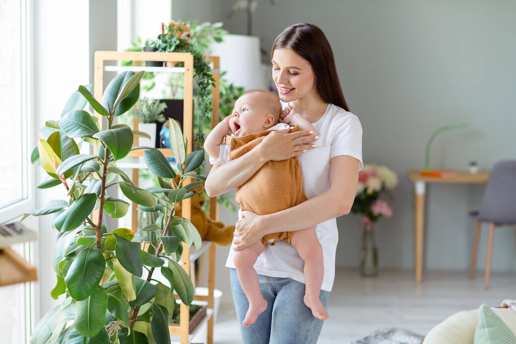Foto de cuidar positivo alegre mãe ruiva senhora segurar gritando bebê desgaste branco t-shirt casa dentro de casa. - Foto, Imagem