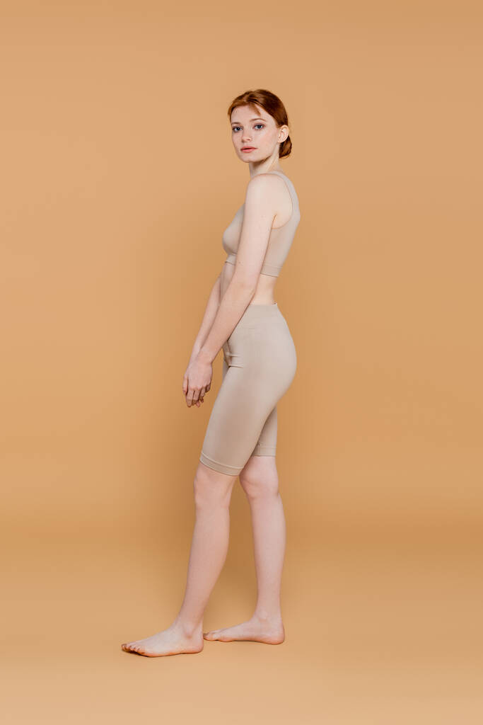 Barefoot red haired woman looking at camera while standing on beige background  - Photo, Image