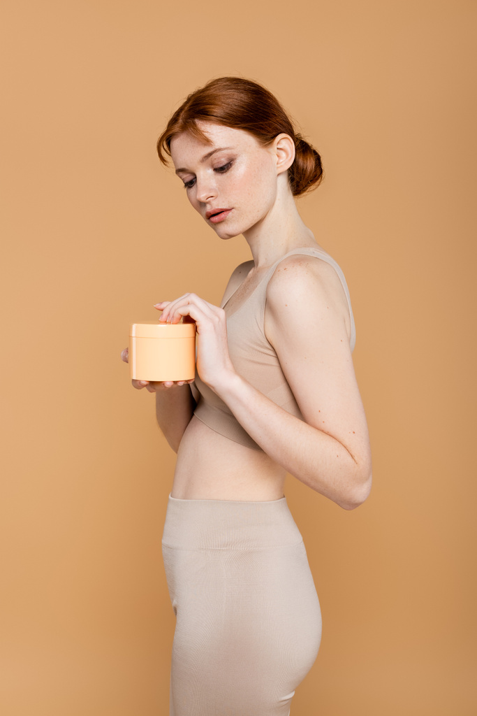 Mujer pecosa y pelirroja sosteniendo crema cosmética en recipiente aislado en beige  - Foto, imagen