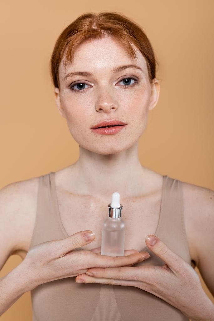 jonge roodharige vrouw met cosmetisch serum en kijken naar camera geïsoleerd op beige  - Foto, afbeelding