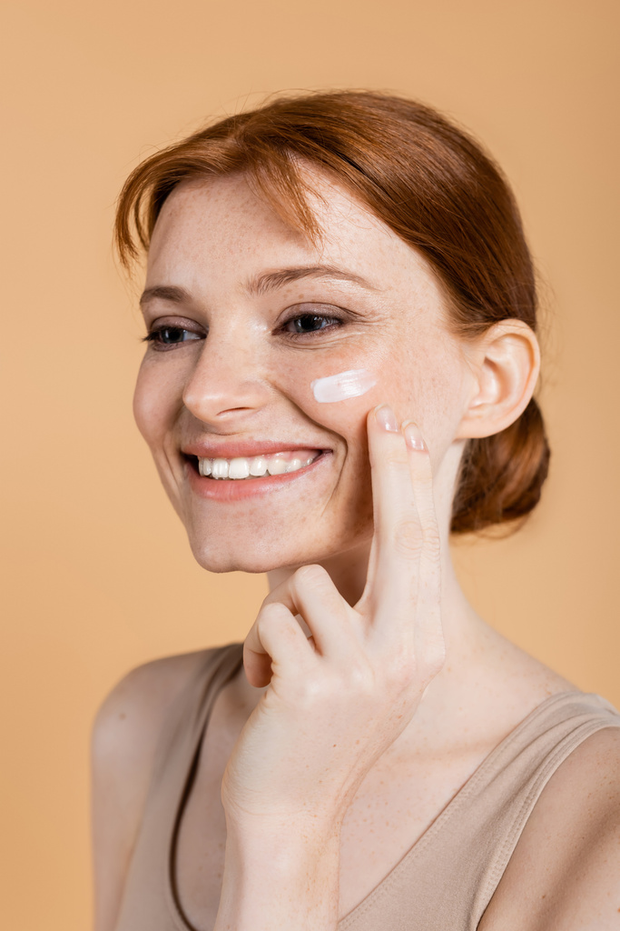Pozitivní červené vlasy žena nanášení krém na tvář izolované na béžové  - Fotografie, Obrázek