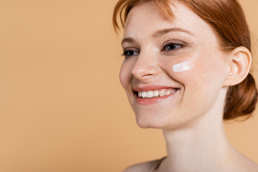 Femme rousse souriante avec peau parfaite et crème sur le visage regardant loin isolé sur beige  - Photo, image