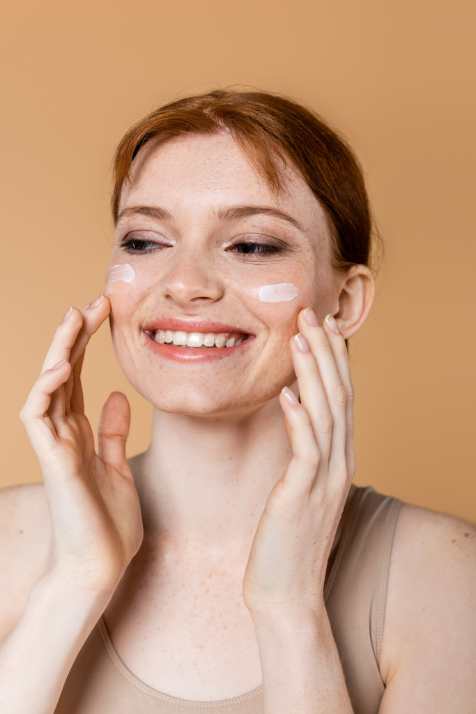 vrolijke rood harige vrouw aanbrengen cosmetische crème op wangen geïsoleerd op beige  - Foto, afbeelding