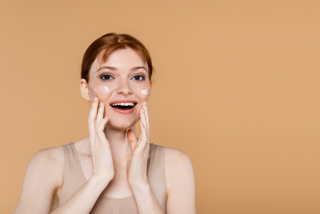 Excited red haired woman with cosmetic cream touching cheeks isolated on beige  - Photo, Image