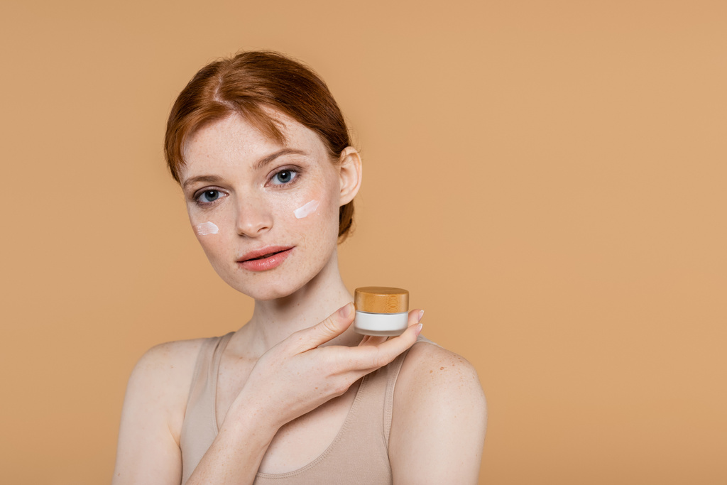 Bella giovane donna con crema sul viso tenuta vaso isolato su beige  - Foto, immagini