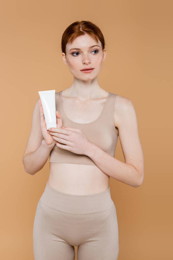 Jolie femme aux cheveux rouges tenant tube avec crème cosmétique isolée sur beige  - Photo, image