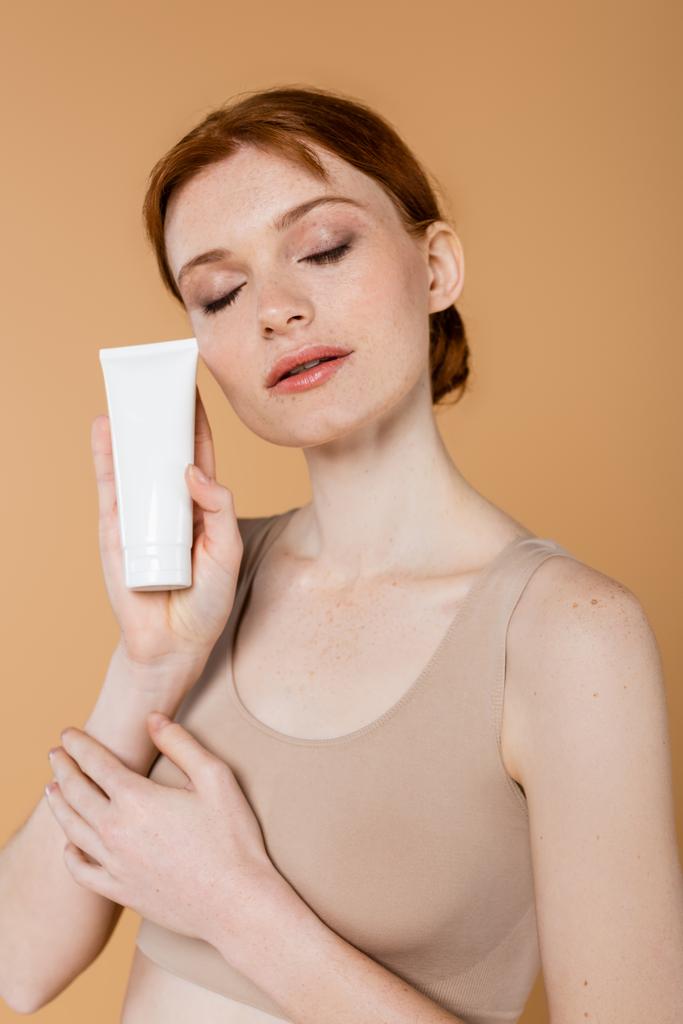 Mujer joven pelirroja con los ojos cerrados sosteniendo tubo con loción cosmética aislada en beige  - Foto, imagen