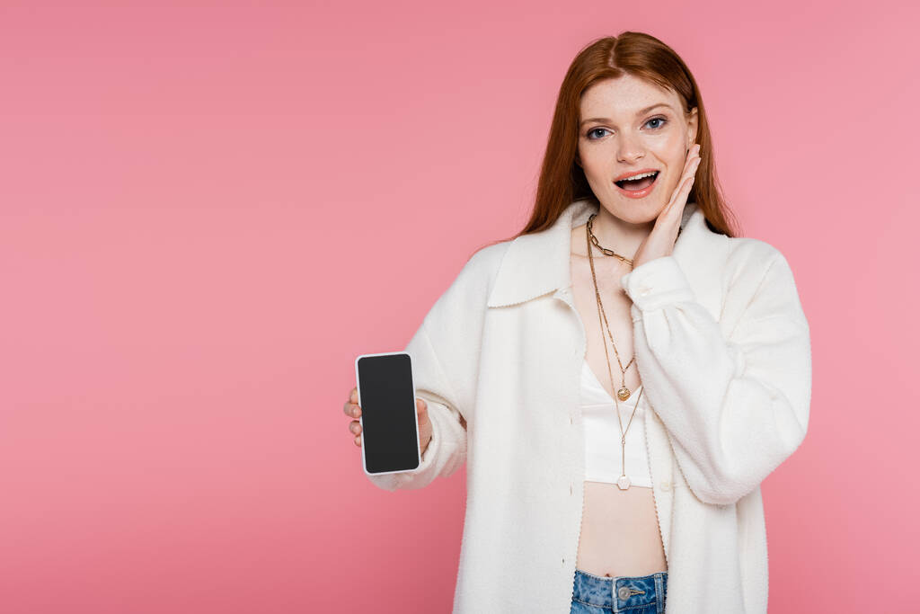 Nadšený ryšavý žena ukazuje smartphone s prázdnou obrazovkou izolované na růžové - Fotografie, Obrázek