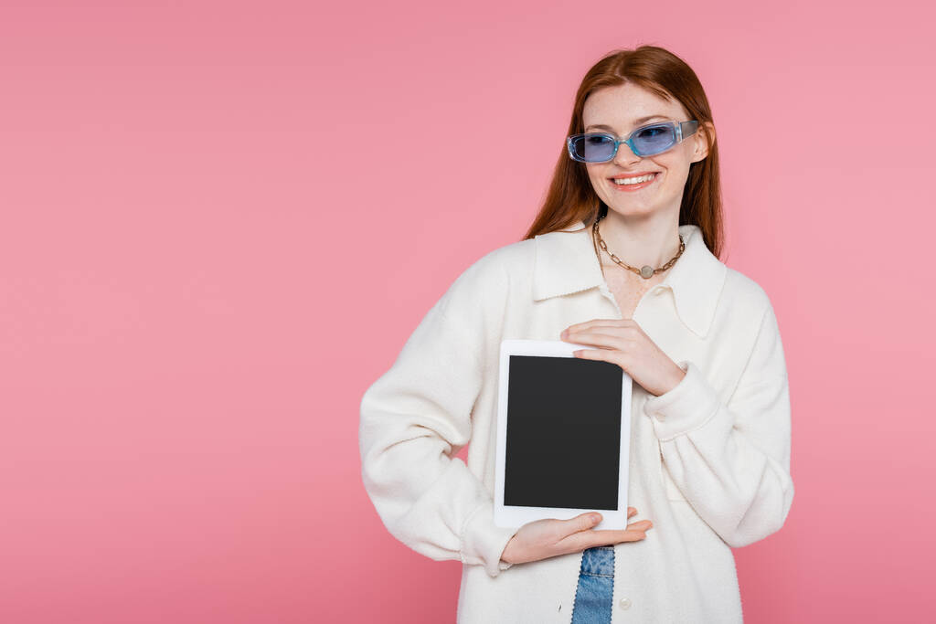 Csinos vörös hajú nő dzsekiben és napszemüvegben digitális tablettát tart üres képernyővel elszigetelt rózsaszín - Fotó, kép