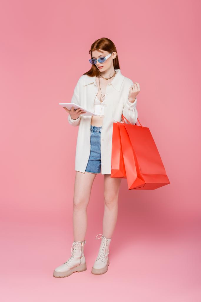 Elegante mujer pelirroja sosteniendo tableta digital y bolsas de compras sobre fondo rosa - Foto, imagen