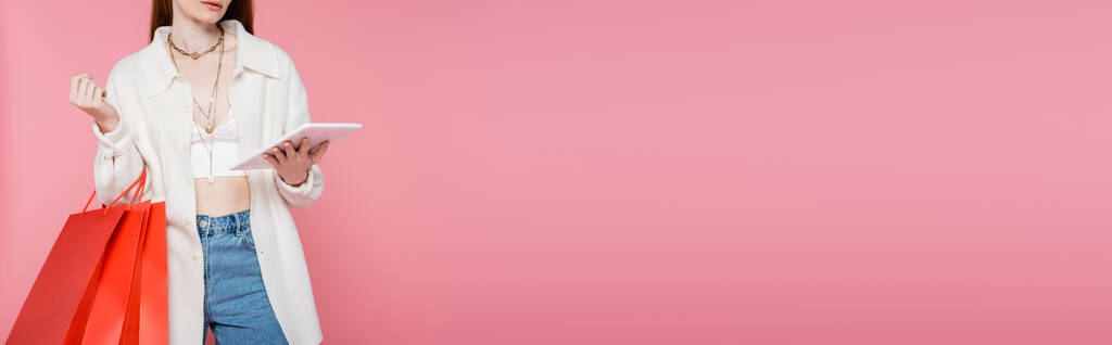 Vista recortada de la mujer elegante sosteniendo bolsas de compras y el uso de tableta digital aislado en rosa, bandera  - Foto, Imagen