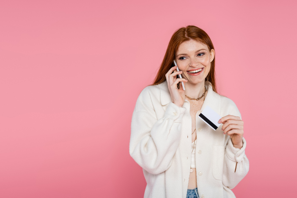 Elegante mujer pecosa hablando en el teléfono inteligente y la celebración de la tarjeta de crédito aislado en rosa  - Foto, Imagen