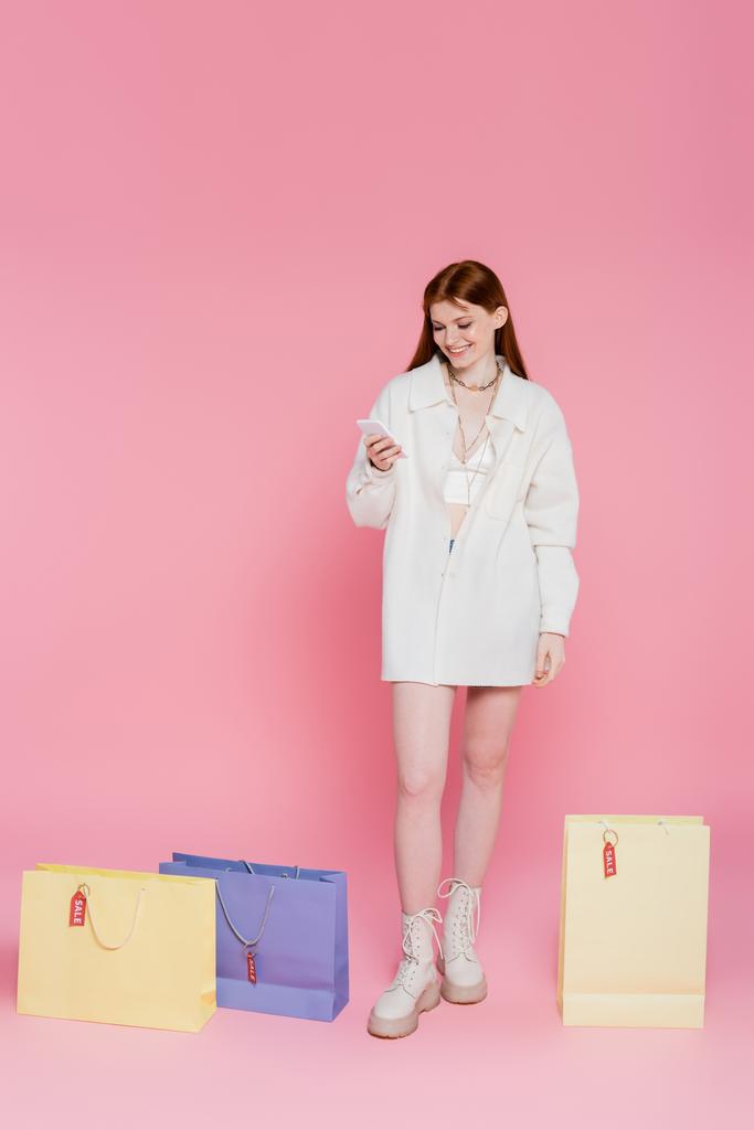 Smiling shopaholic usando smartphone cerca de bolsas de compras con etiquetas de precios y letras de venta sobre fondo rosa - Foto, Imagen