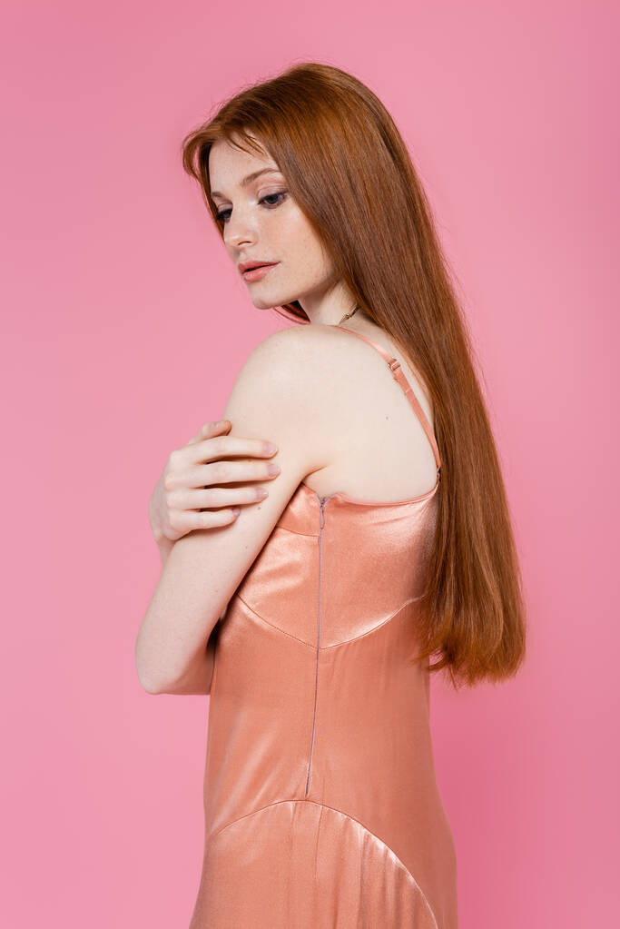 Joven mujer pelirroja en vestido de seda mirando hacia abajo aislado en rosa  - Foto, Imagen