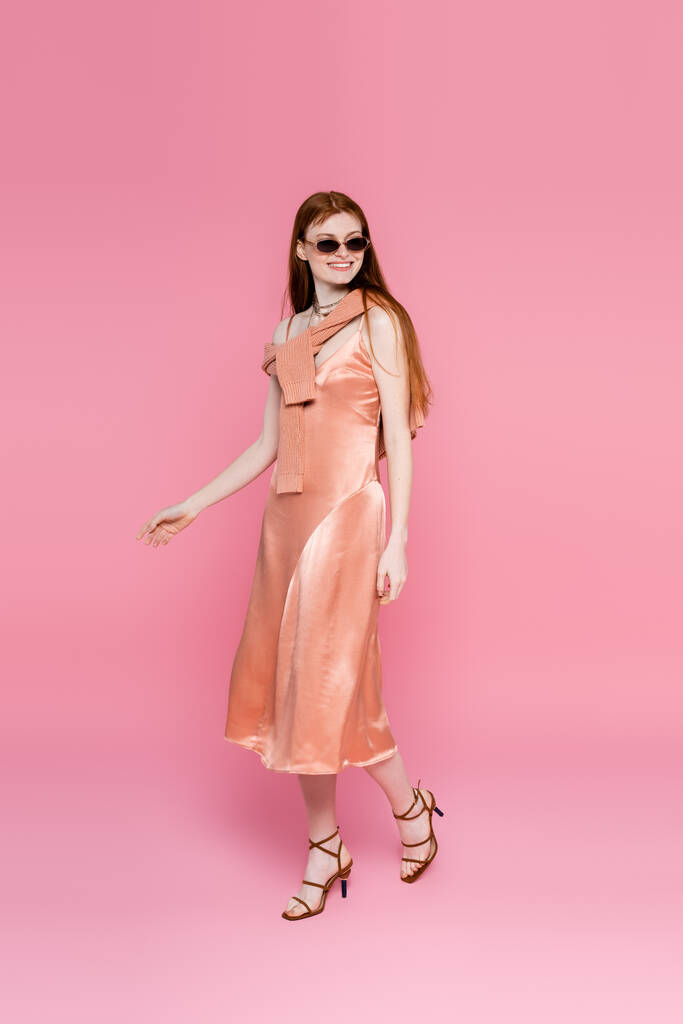 ピンクの背景を歩く絹のドレスとサングラスのスタイリッシュな女性  - 写真・画像