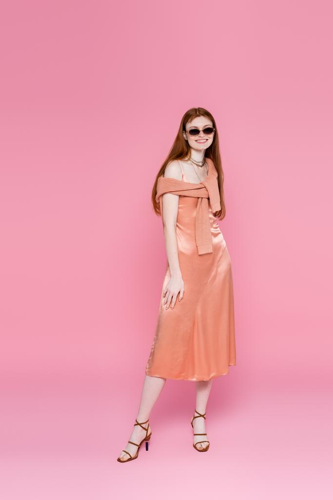 Volle Länge einer lächelnden Frau in Kleid und Sonnenbrille, die auf rosa Hintergrund posiert  - Foto, Bild