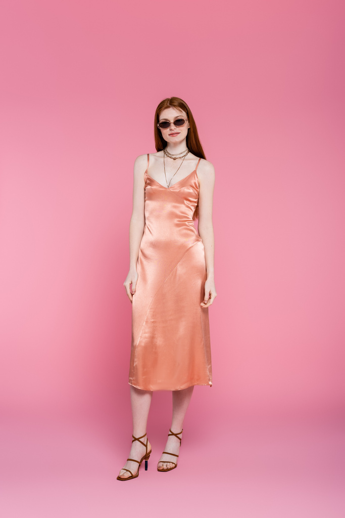 Full length of young red haired woman in sunglasses and dress standing on pink background  - Photo, Image