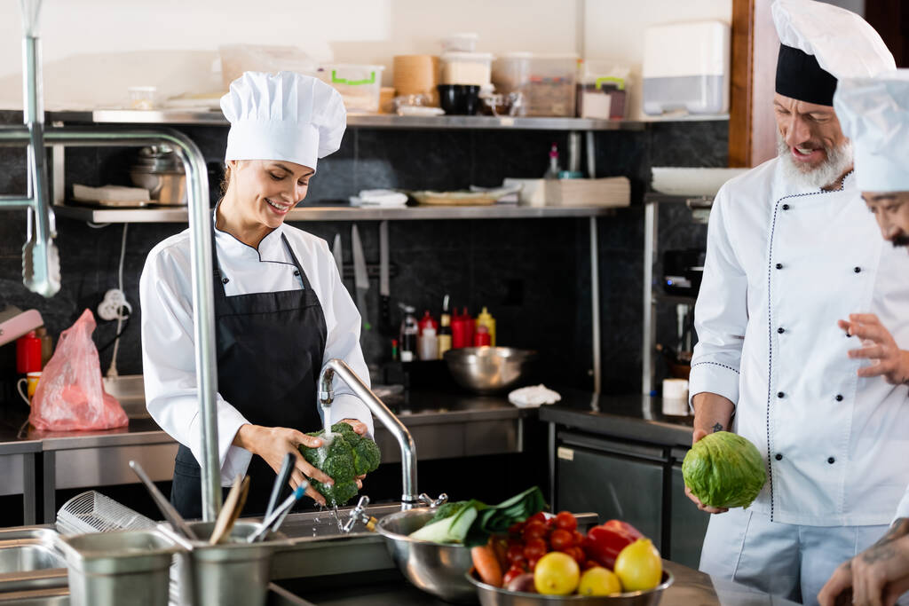 Улыбающийся шеф-повар стирает брокколи рядом с межрасовыми коллегами разговаривая на кухне ресторана  - Фото, изображение