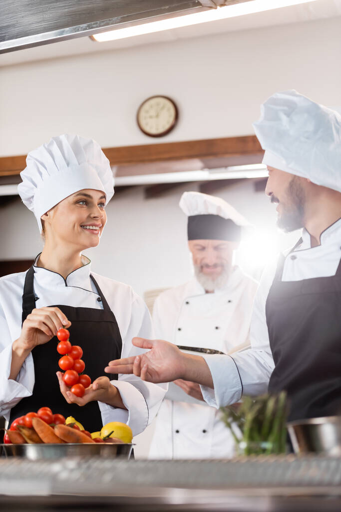 Усміхнений азіатський шеф-кухар вказує на помідори вишні поблизу колеги і стиглі овочі на кухні
  - Фото, зображення