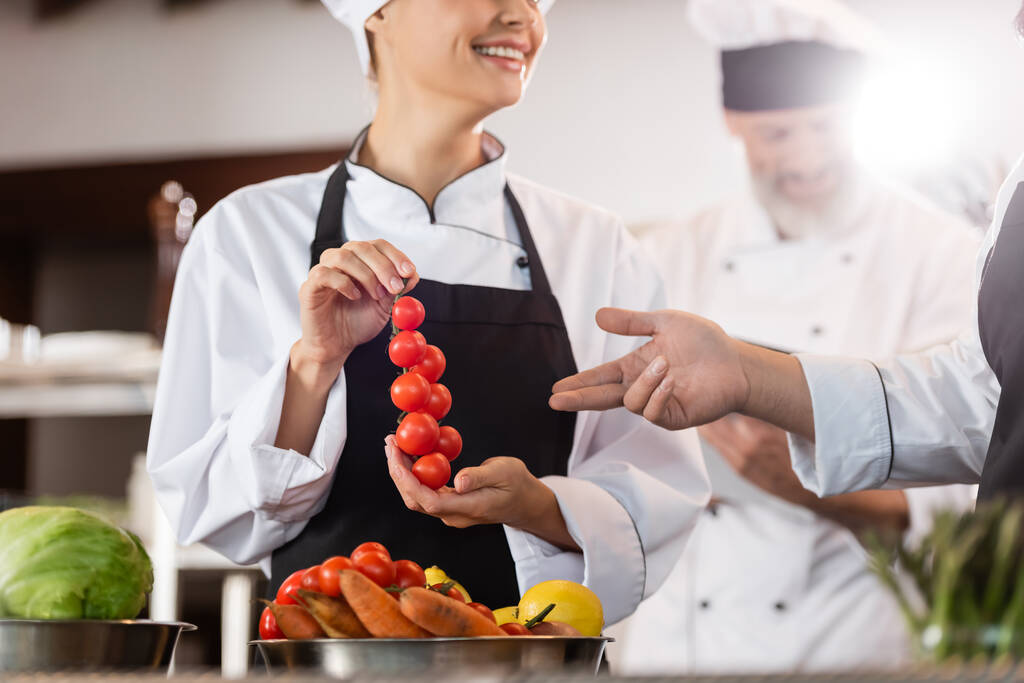 Chef apuntando a los tomates cherry cerca de colega sonriente en la cocina del restaurante  - Foto, Imagen