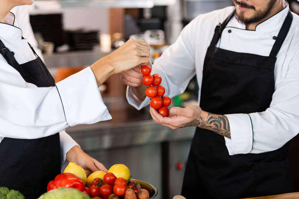 Обрізаний вид шеф-кухарів в фартухах, що тримають вишневі помідори на кухні
  - Фото, зображення