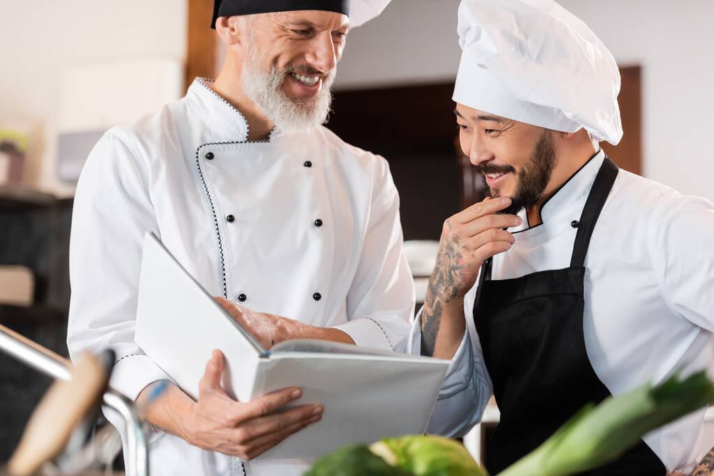 Chefs inter-raciais alegres trabalhando com livro de receitas na cozinha  - Foto, Imagem