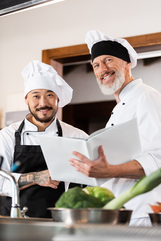 Mature chef holding cookbook near smiling asian colleague and blurred vegetables in kitchen  - Photo, Image