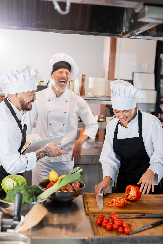 Asian chef holding cookbook near laughing colleagues and fresh food in kitchen  - Photo, Image