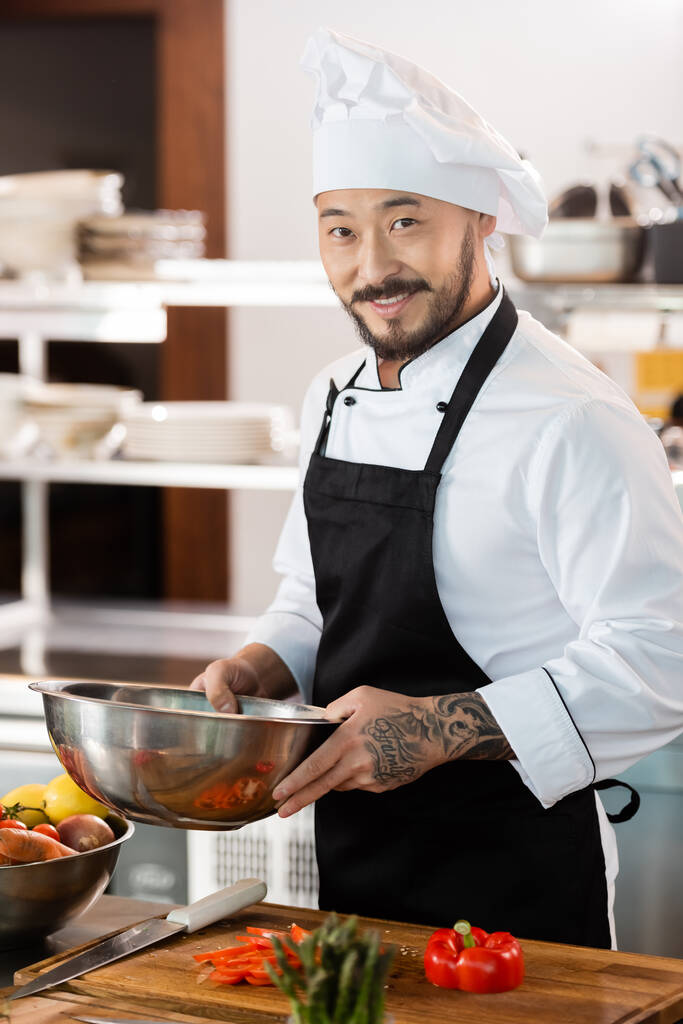 Smiling asian chef in apron holding bowl near cutting board and vegetables in kitchen  - Photo, Image
