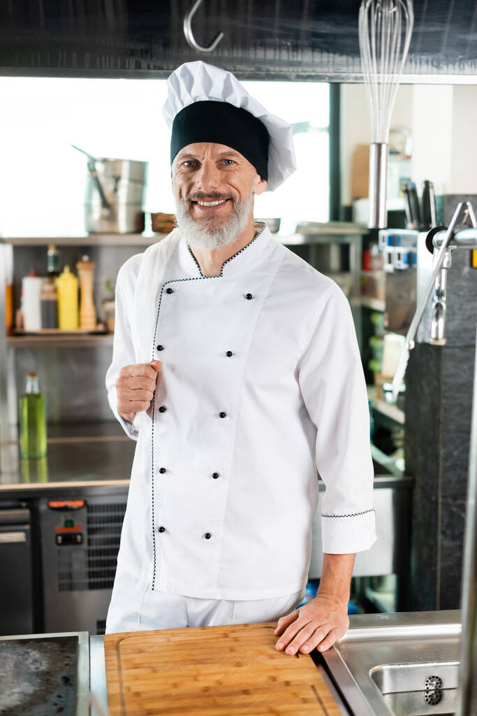 Позитивний шеф-кухар в уніформі дивиться на камеру біля обробної дошки і плити на кухні
  - Фото, зображення