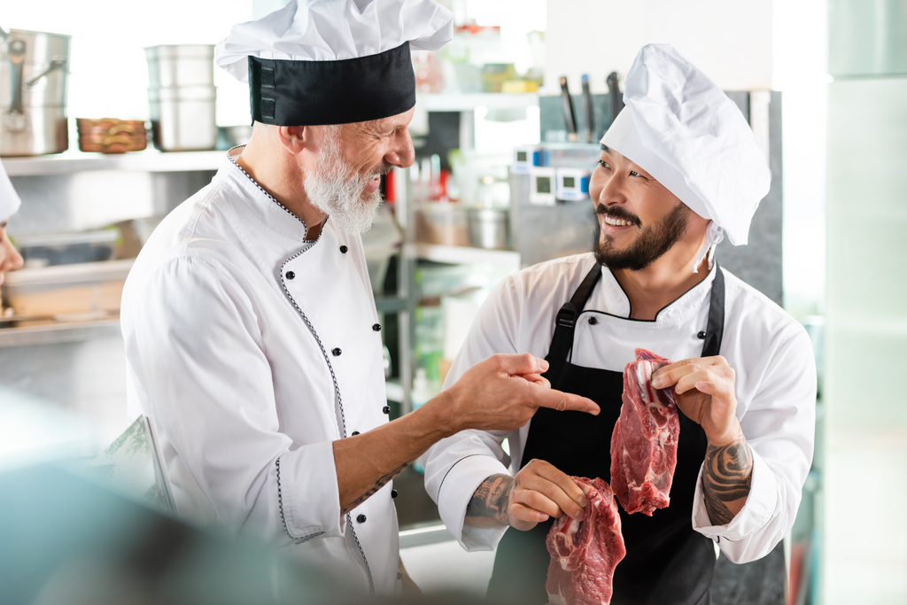 Sonriente chef apuntando a la carne cruda cerca tatuado asiático colega en cocina  - Foto, Imagen