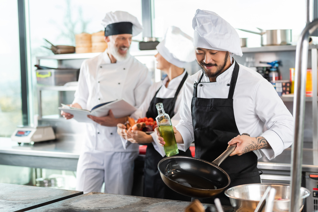 Sonriente asiático chef sosteniendo sartén y aceite de oliva cerca de cooktop y borrosa colegas en la cocina  - Foto, imagen