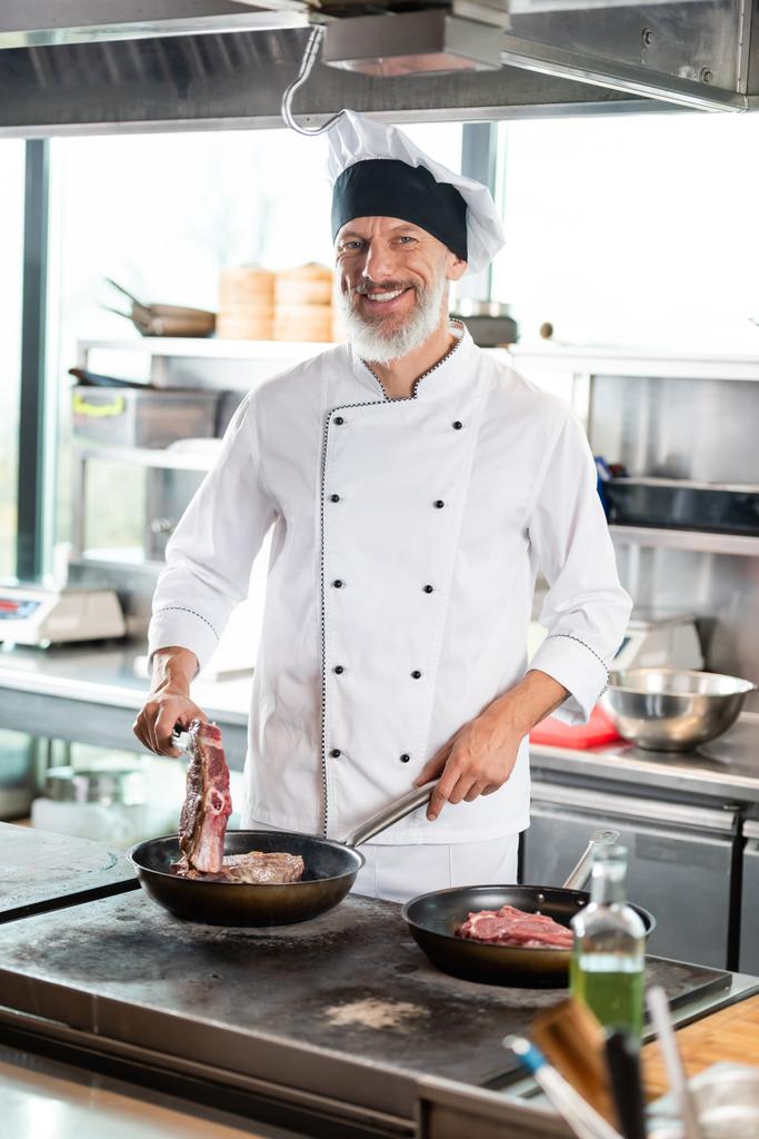 Lächelnder Koch in Uniform kocht Fleisch auf Herdplatte in Restaurantküche  - Foto, Bild