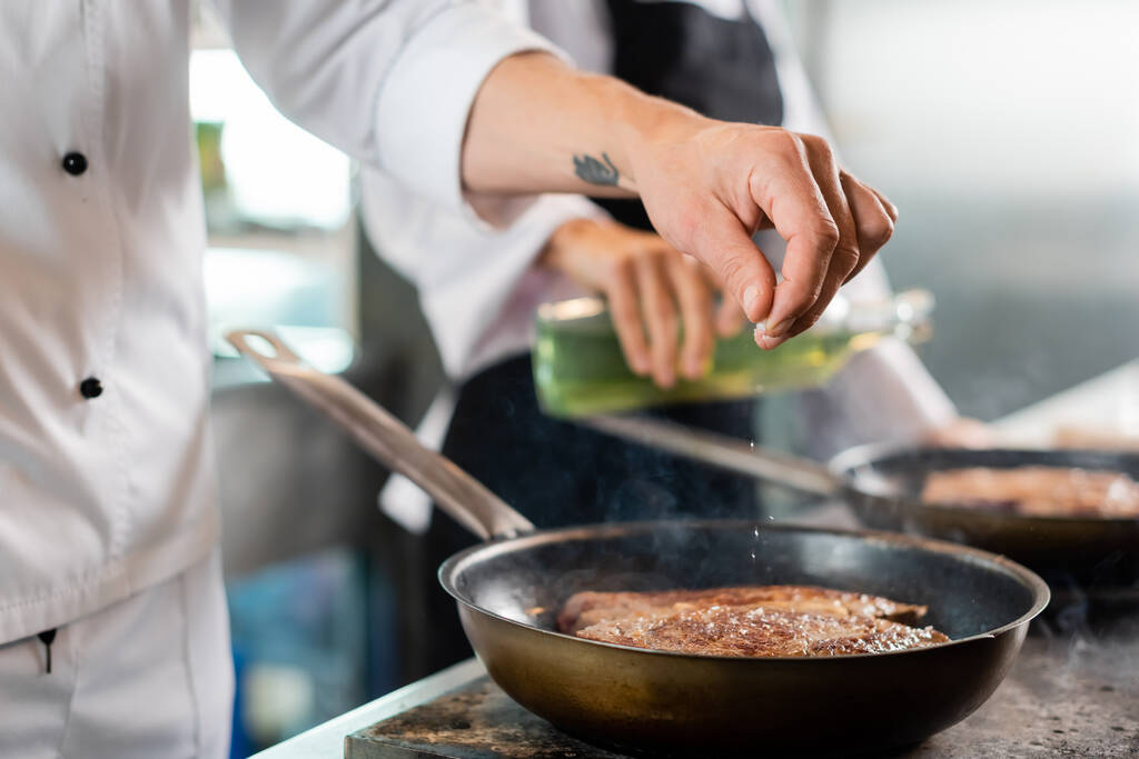 Καλλιεργημένη θέα του σεφ ρίχνει αλάτι, ενώ το μαγείρεμα του κρέατος στο τηγάνι στην κουζίνα  - Φωτογραφία, εικόνα
