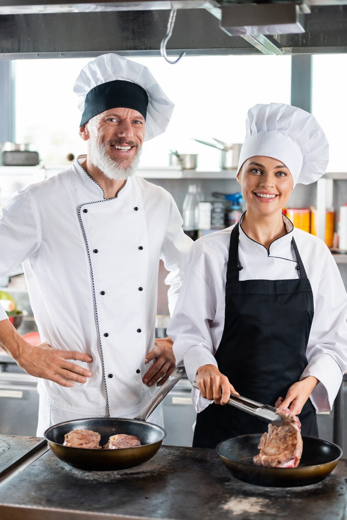 Glimlachende koks in uniform kijkend naar de camera terwijl ze vlees koken in de keuken  - Foto, afbeelding