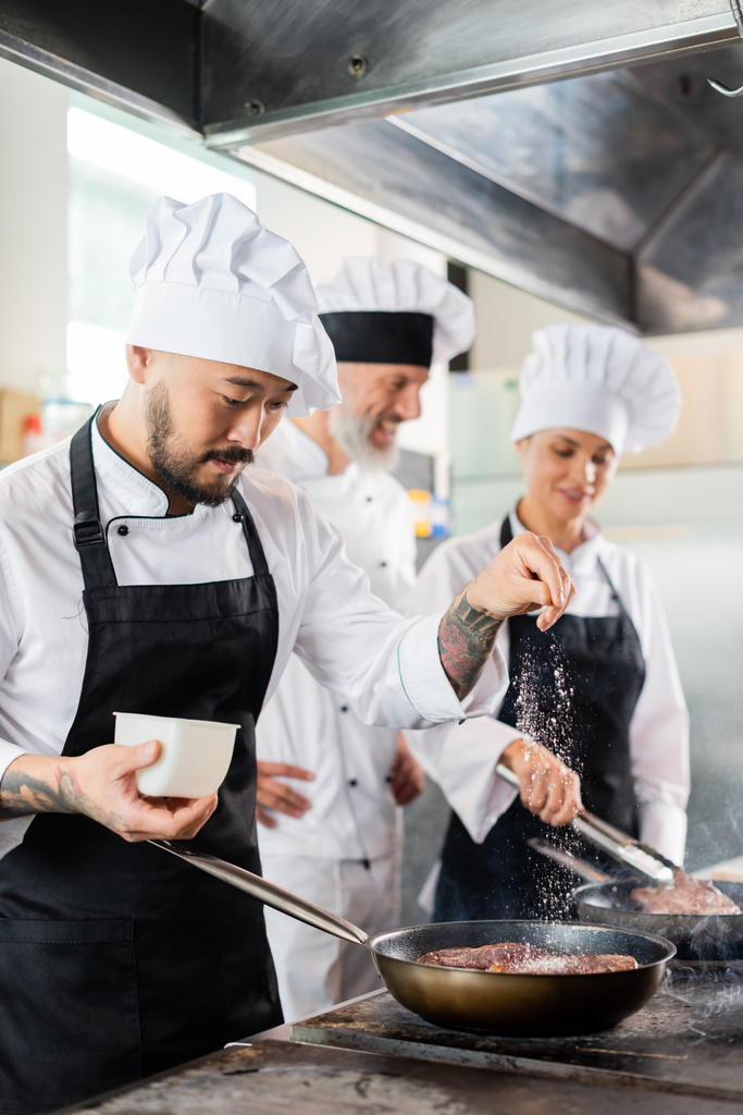 Азиатский шеф-повар льет соль на мясо на плиту рядом с размытыми коллегами на кухне  - Фото, изображение