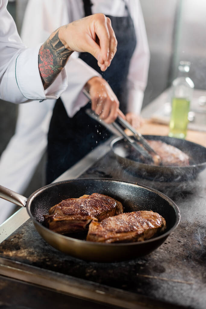 Rajatut näkymät tatuoitu kokki kaatamalla suolaa paahtaessaan lihaa keittotasolla keittiössä  - Valokuva, kuva