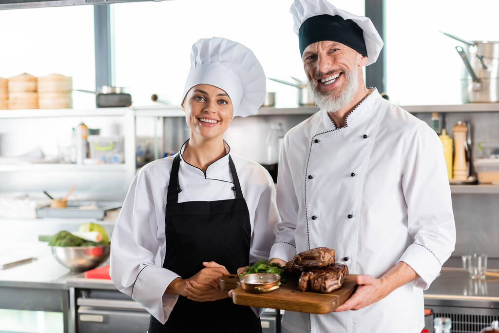 chefs positivos sorrindo para a câmera enquanto segurando carne assada na cozinha  - Foto, Imagem