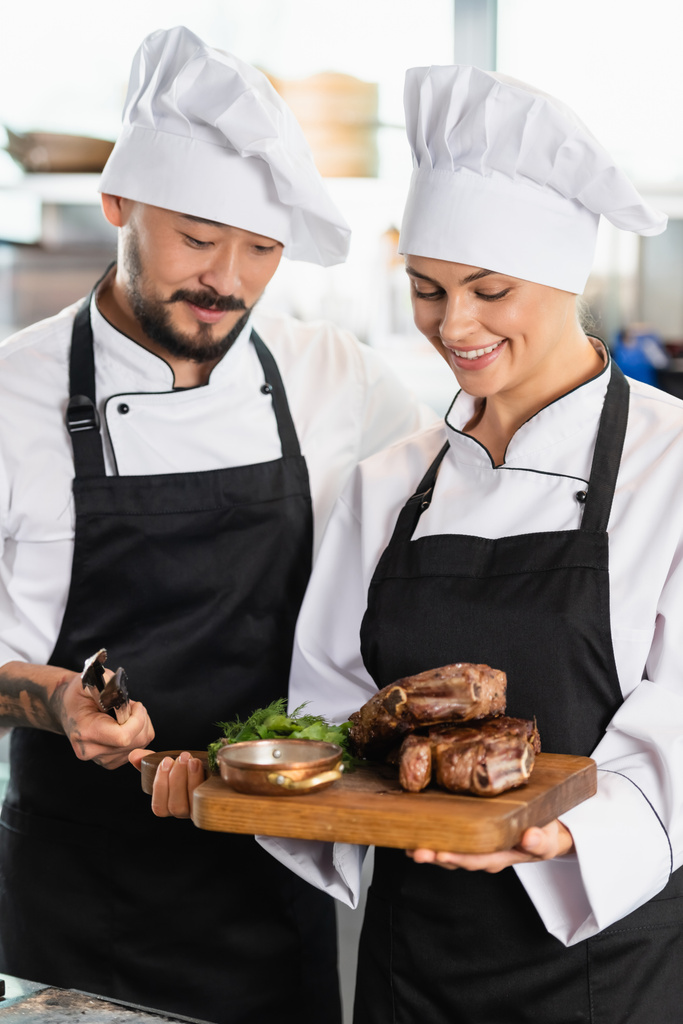Ασιάτης σεφ κρατώντας λαβίδες κοντά χαρούμενος συνάδελφος με ψητό κρέας στον πίνακα κοπής  - Φωτογραφία, εικόνα