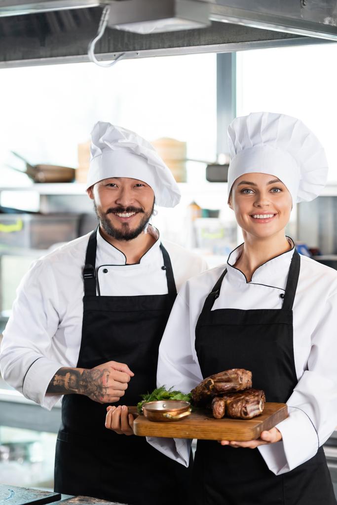 Татуированный азиатский шеф-повар, стоящий рядом с коллегой с жареным мясом на разделочной доске на кухне  - Фото, изображение