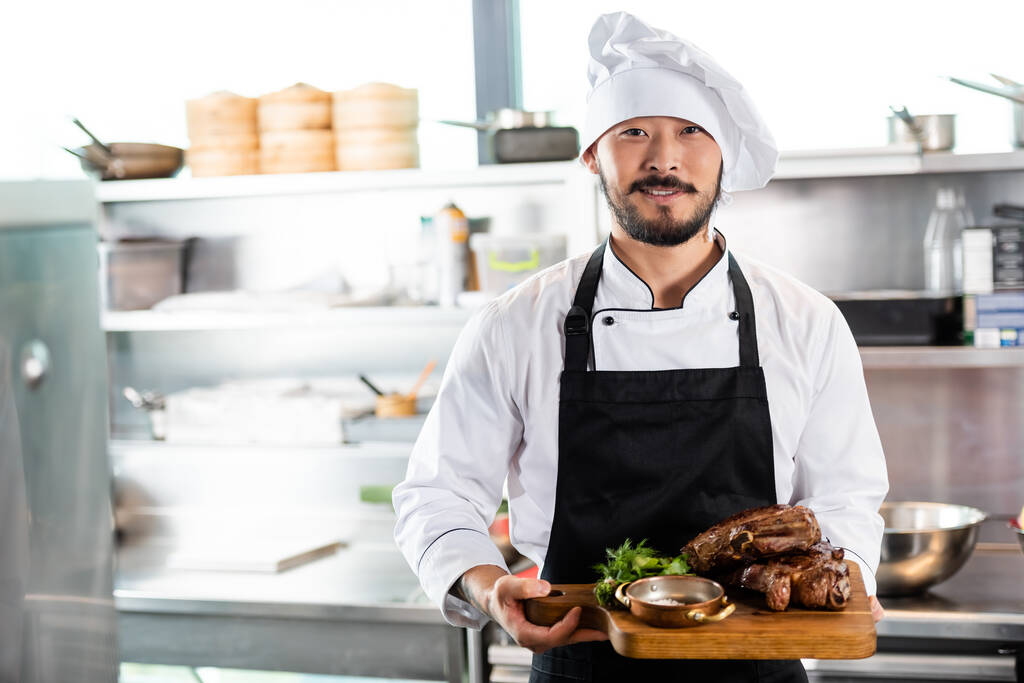Sorrindo asiático chef segurando tábua de corte com carne assada e verduras na cozinha  - Foto, Imagem