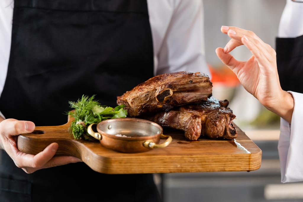 Καλλιεργημένη άποψη του σεφ δείχνει εντάξει χειρονομία κοντά συνάδελφο με ψητό κρέας στον πίνακα κοπής  - Φωτογραφία, εικόνα
