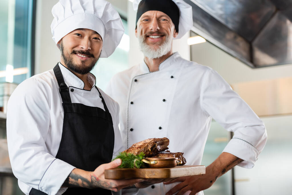 Sorrindo asiático chef segurando placa de corte com carne assada perto colega na cozinha  - Foto, Imagem