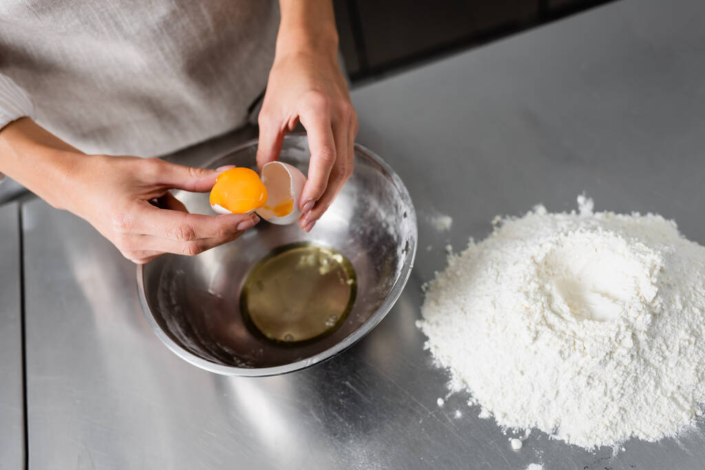 Vista recortada del chef sosteniendo huevo y cáscara de huevo cerca del tazón y harina en la mesa  - Foto, Imagen
